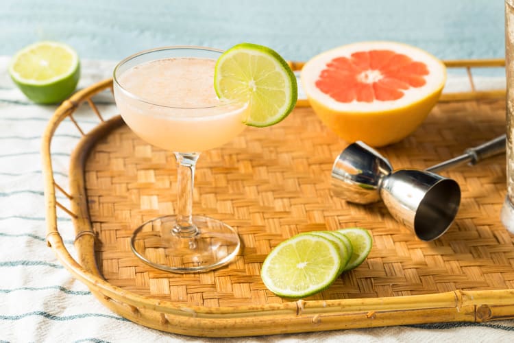 A daiquiri, a popular rum cocktail, on a serving tray with lemon wedges, half a grapefruit and a shot pourer