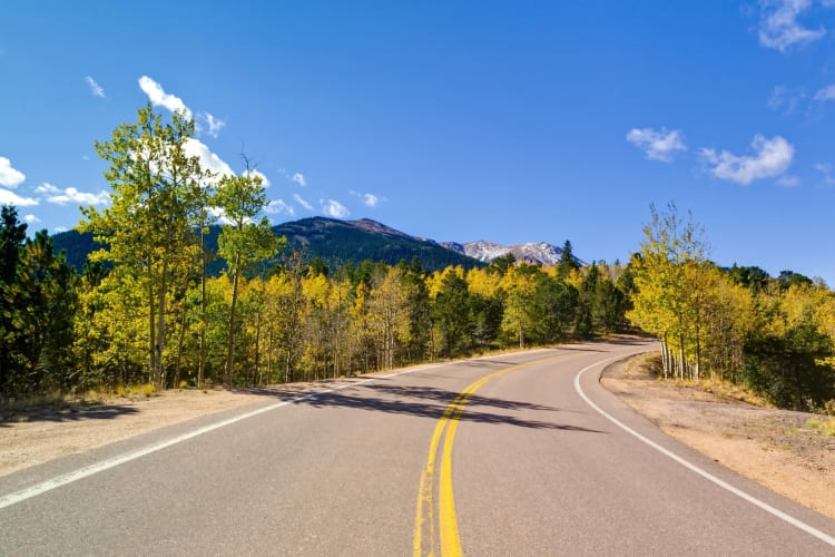 A road through green trees next to mountains under a blue sky