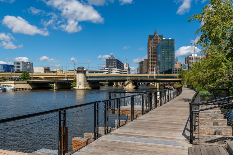 A boardwalk next to a river, trees and city buildings