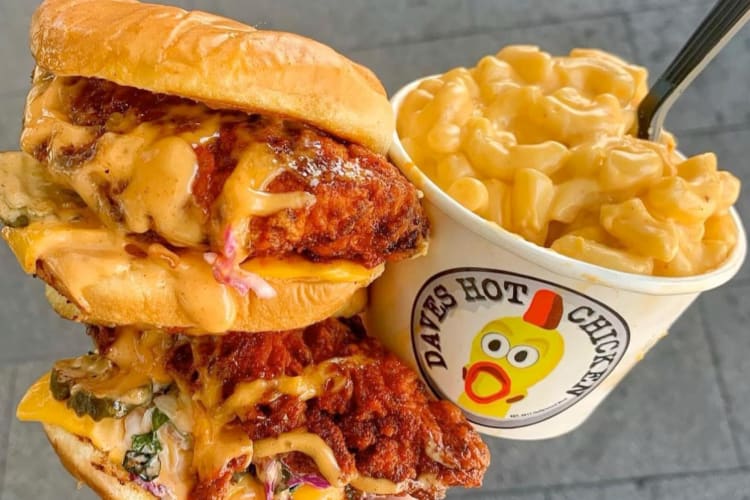 A person holding two hot chicken sandwiches and a tub of mac and cheese