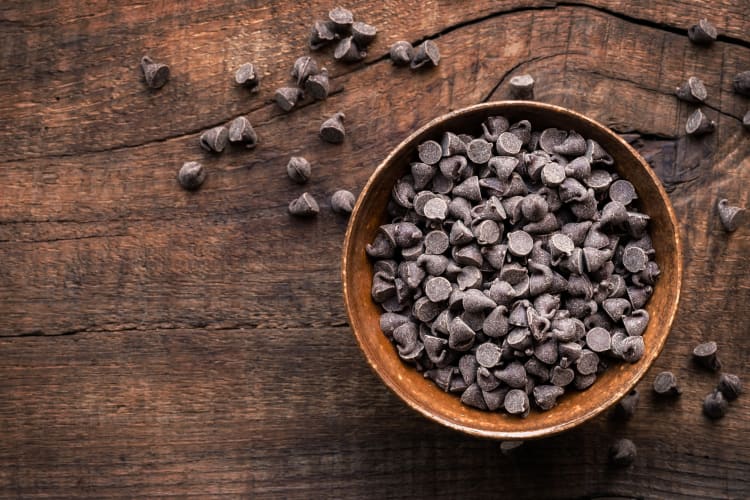 A bowl of chocolate chips with more scattered around it on a wooden surface