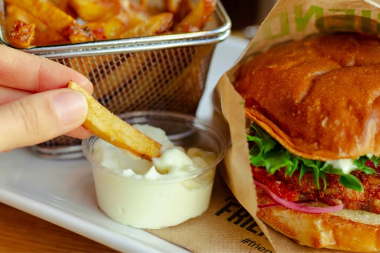 A person dipping a French fry in mayonnaise next to a burger