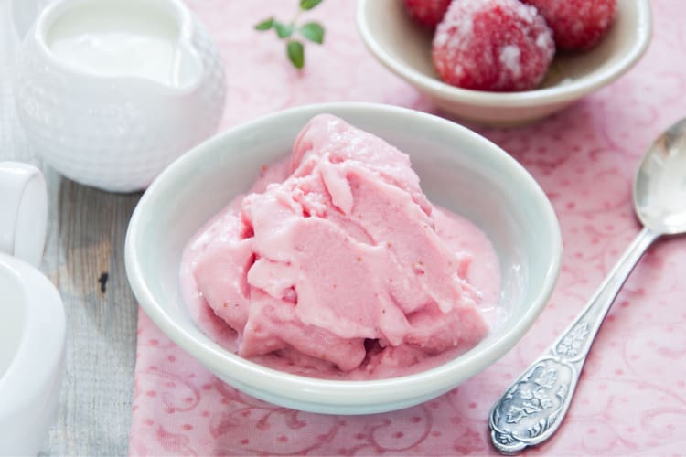 A bowl of pink frozen yogurt next to a spoon, small milk jugs and fruit