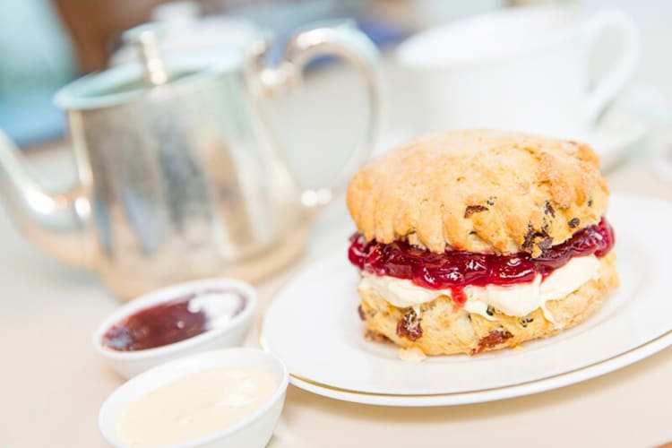 A scone with cream and jam next to a silver teapot