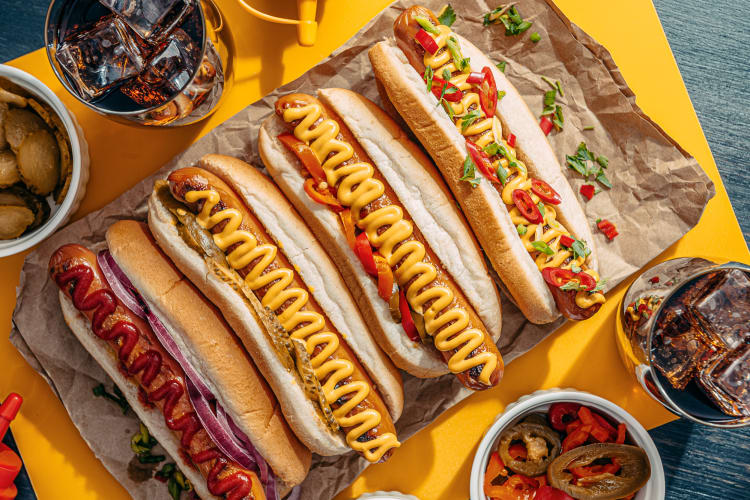 Four hot dogs with different toppings, surrounded by more toppings and drinks