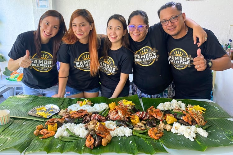 Five people standing in front of a table covered in leaves with food on top of them, at the Famous Food Festival