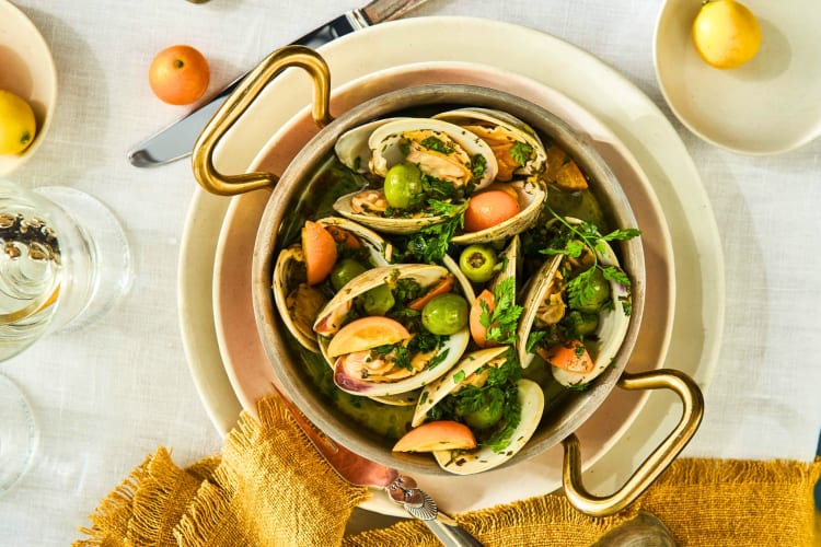 A clam and olive dish on a table with a yellow cloth and citrus fruits