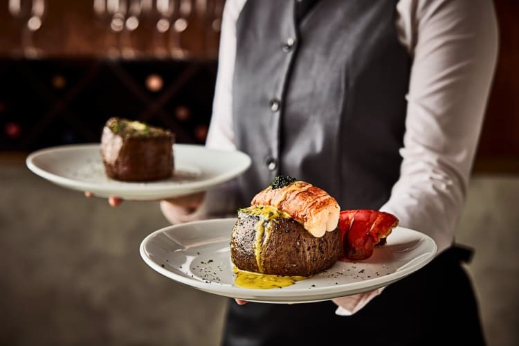 A waiter holding two plates with a steak on each, from Fleming's Prime Steakhouse & Wine Bar