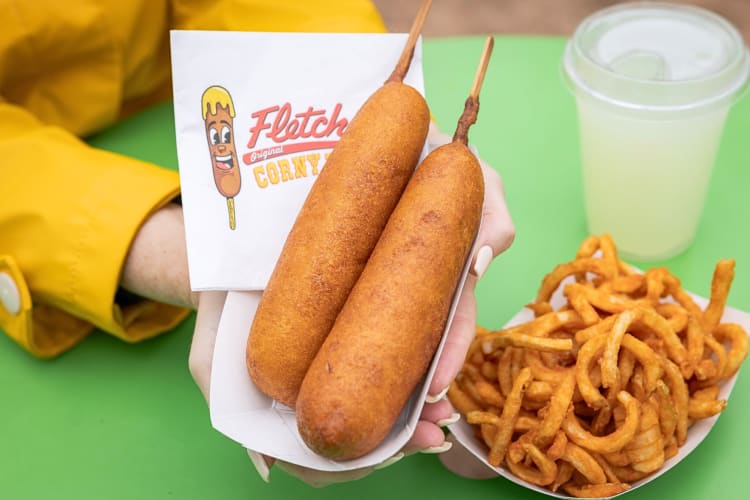 Person's hands holding two corn dogs next to curly fries, from Fletcher’s Original Corny Dogs