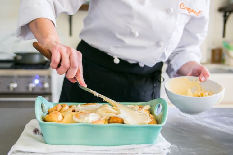 A person in a Cozymeal apron putting frosting on cinnamon rolls