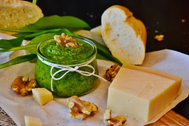 A jar of pesto next to walnuts, cheese and olive oil