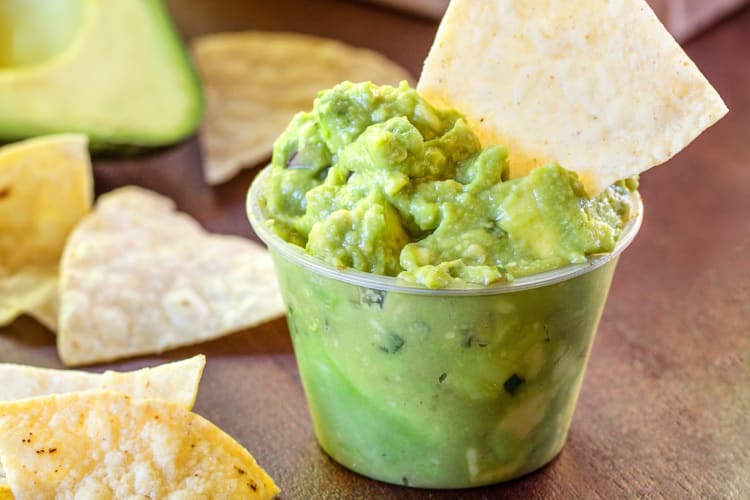 Tupperware of guacamole with a chip in it and chips surrounding it