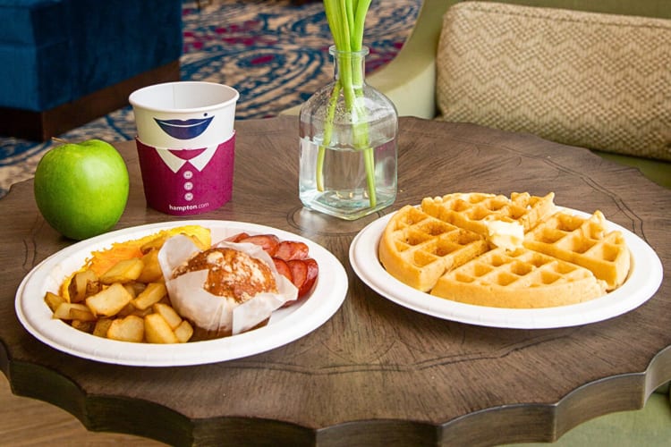 Two breakfast plates, one with waffles and one with a muffin, sausages, hash browns and potatoes