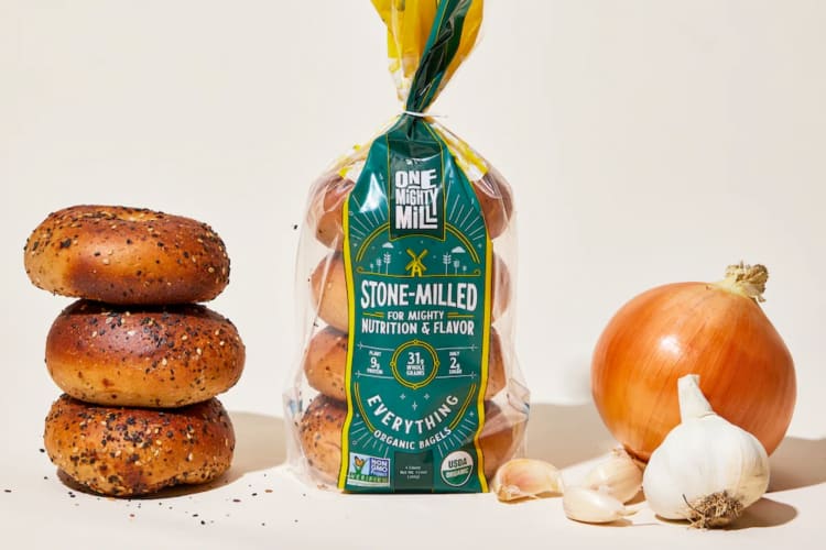 A bag of bagels next to a stack of bagels and fresh onion and garlic