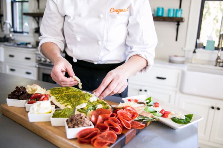 A person in a Cozymeal apron preparing a charcuterie board