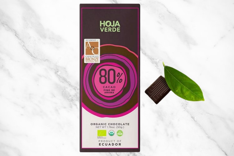 A chocolate bar next to a piece of chocolate and a leaf