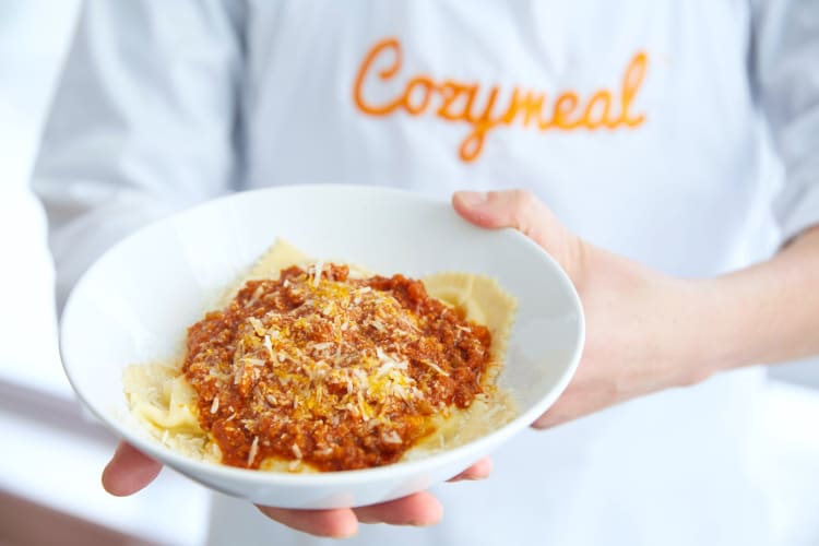 A person in a Cozymeal apron holding a bowl of pasta with ragu sauce