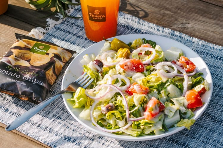 A salad in a bowl next to a bag of potato chips and a drink