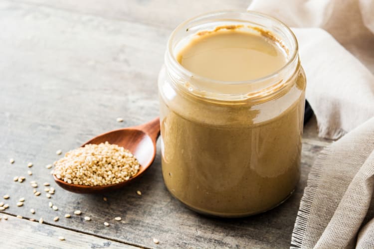 A jar of tahini next to a wooden spoon with sesame seeds