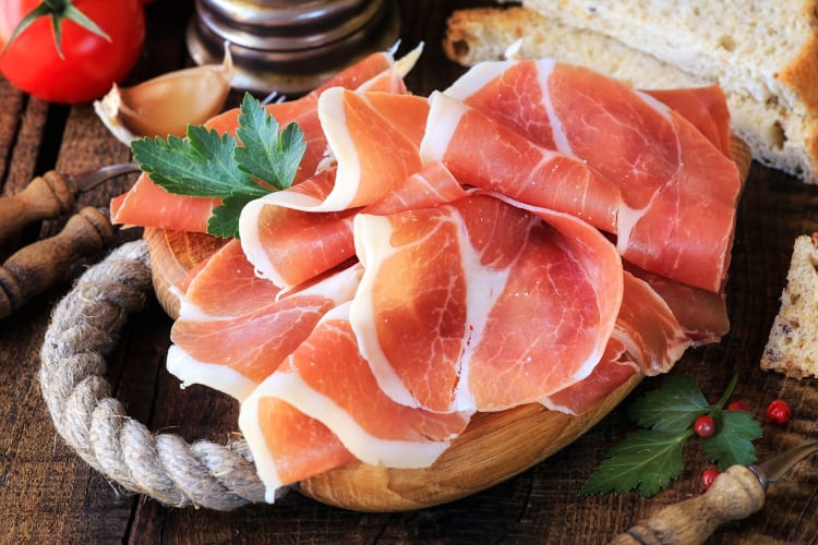 Pieces of prosciutto on a wooden chopping board next to bread and a tomato