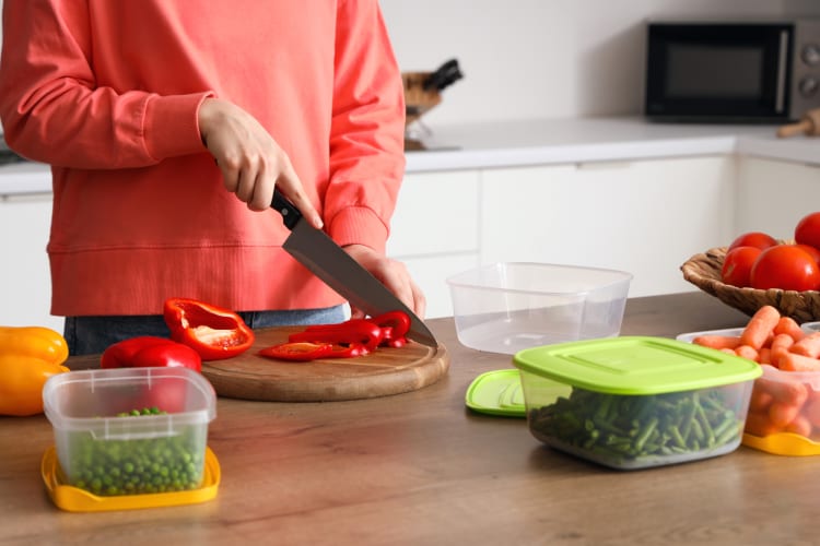 Person cutting a red bell pepper in the background, with other vegetables in the foreground