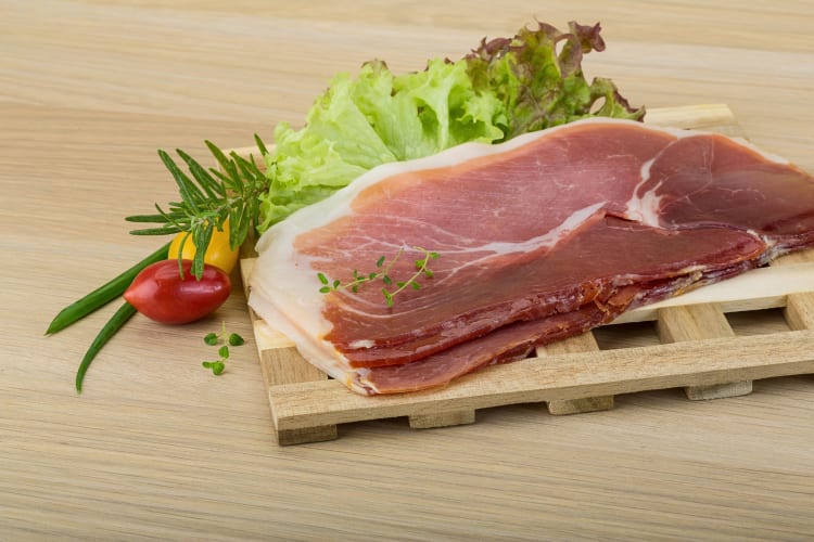 Prosciutto slices next to greens on a wooden chopping board