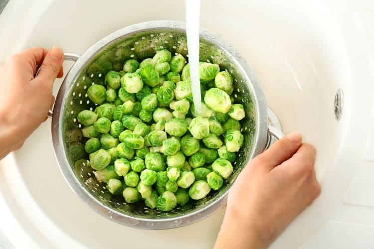 hands holding a strainer with Brussels sprouts in it being washed under water