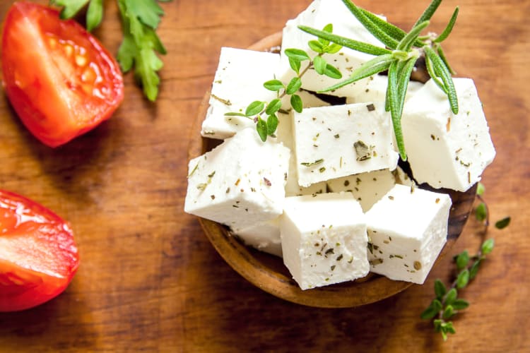 Blocks of feta cheese with herbs in a bowl next to tomatoes