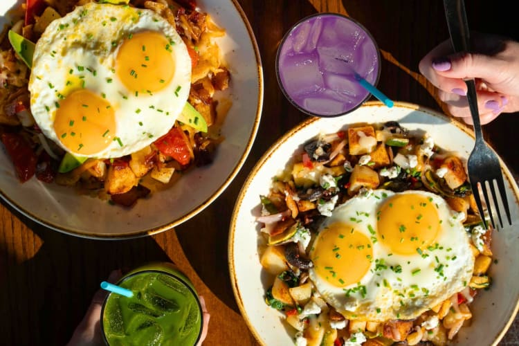 Two breakfast hash dishes next to green and purple drinks on a table