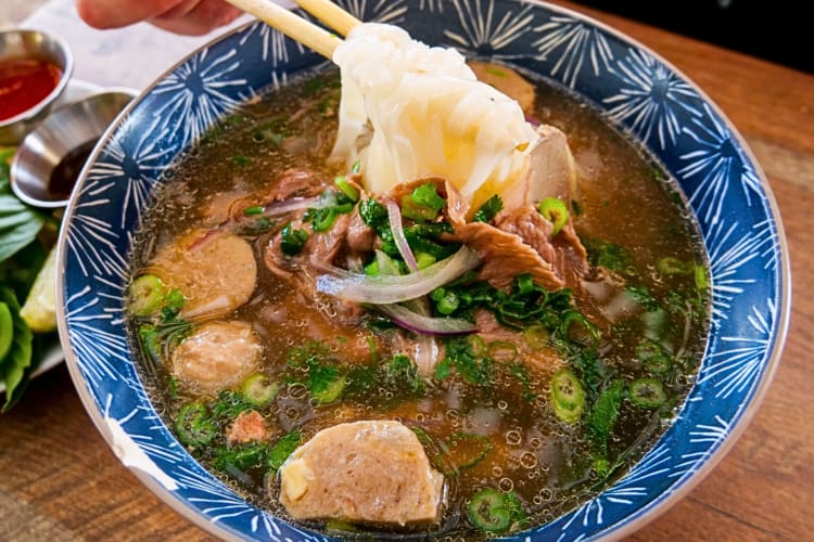 Chopsticks lifting food from a bowl of pho, from Kâu Ba