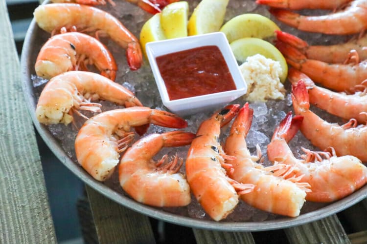 A plate of pink shrimp on ice with lemon and a bowl of sauce