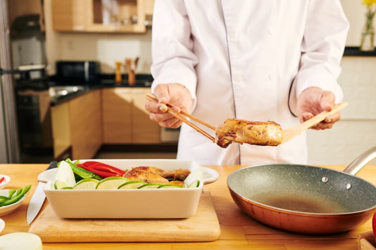 Chef moving a piece of chicken from a pan to a dish with more chicken and greens