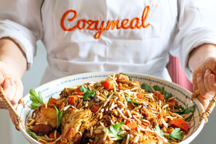A chef in a Cozymeal apron presents a delicious, colorful dish