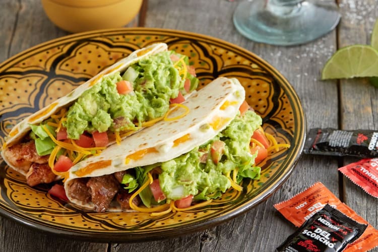 Tacos with guacamole on a yellow and black plate next to sauce packets