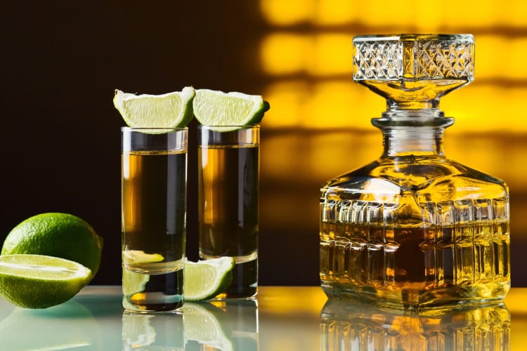 A glass bottle of tequila next to shot glasses and lime