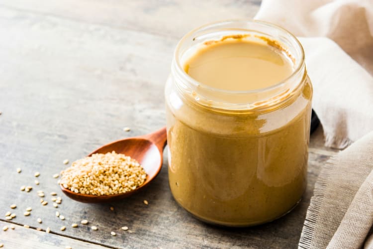 A jar of tahini next to sesame seeds in a wooden spoon