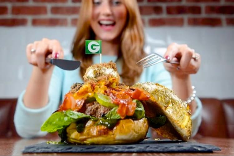 A woman with a knife and fork next to a burger with lots of toppings