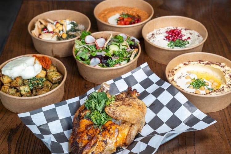 A roast chicken next to potatoes, salad and hummus in containers