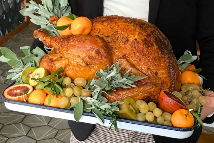 A person holding a roast turkey surrounded by fresh fruit
