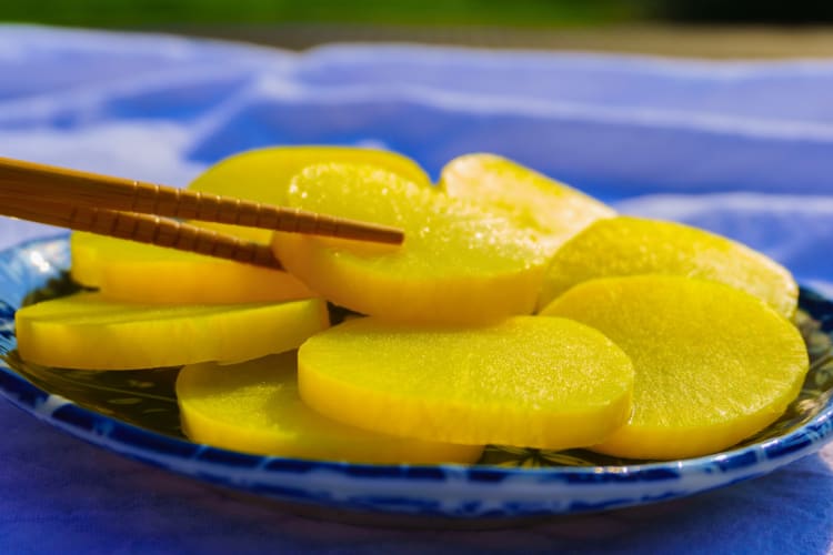 Yellow oshinko in a blue bowl with one piece being picked up by chopsticks