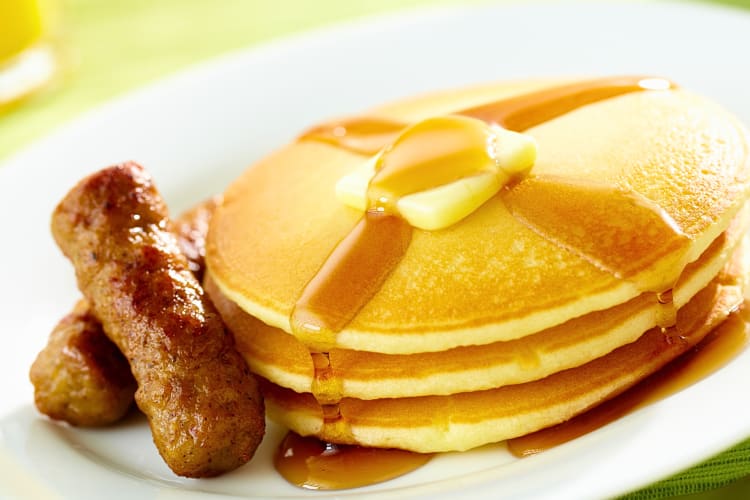 Three pancakes topped with syrup and butter next to two sausages