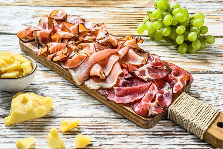Ham, prosciutto, pancetta and bacon slices on a board, next to grapes and pineapple