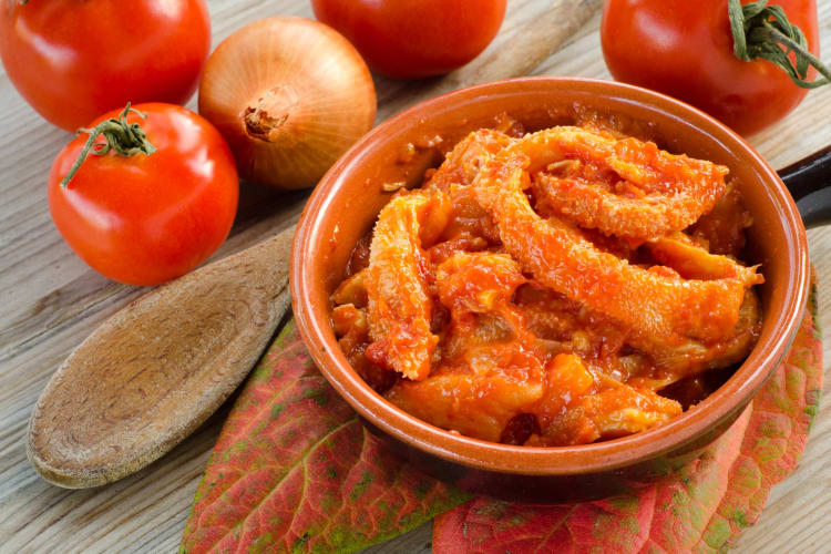 A bowl of tomato and tripe stew next to fresh tomatoes and onions