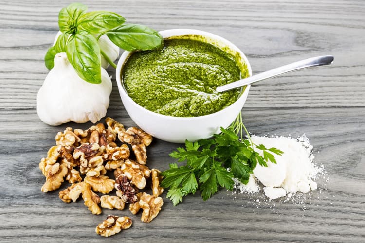 Pesto in a bowl with a spoon, surrounded by walnuts, basil, parsley and garlic