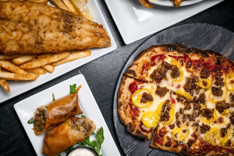 A plate of fish & chips, pizza and spring rolls on a table.