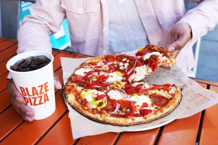 A person taking a slice of pizza and holding a soda cup