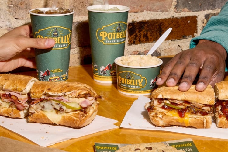 Two people at a table with sandwiches, drinks and other Potbelly dishes