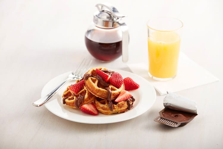 Waffles topped with strawberries and chocolate sauce, next to a glass of juice and jar of syrup