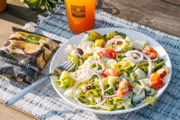 A salad, bag of chips and orange drink on a picnic rug
