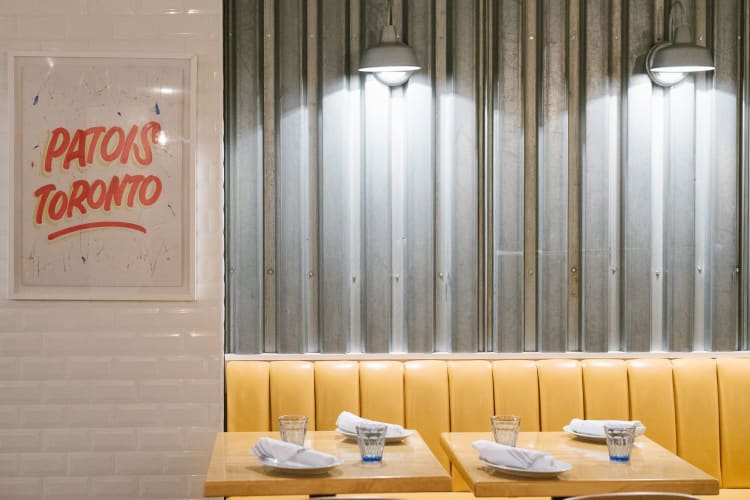 Yellow booth seating tables in front of an industrial metal wall with a sign to the left reading 'Patois Toronto'.